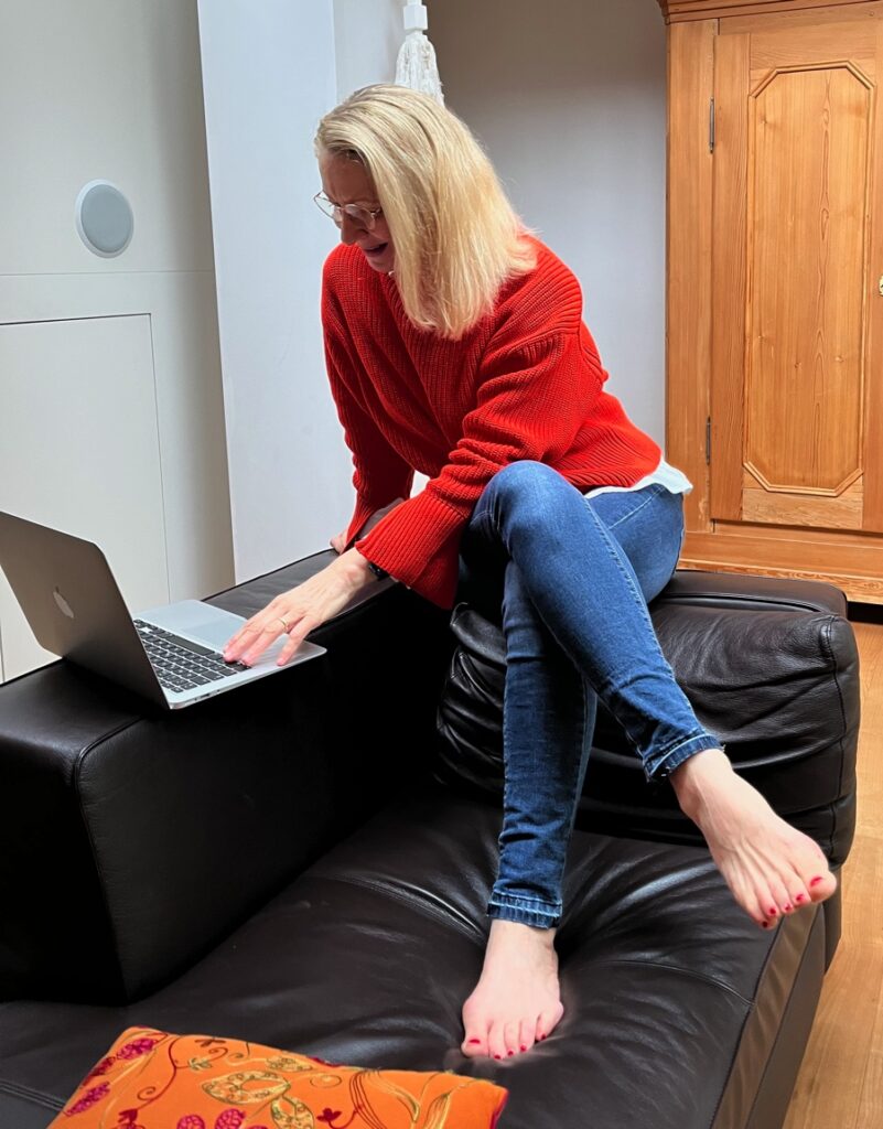 Blonde Frau in orangem Pullover und blauen Jeans sitzt lässig auf Sofarand und schaut in Laptop.