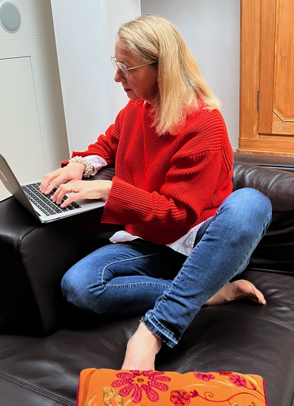 Blonde Frau in orangen Pullover und Jeans sitzt auf braunem Sofa und schreibt am Laptop